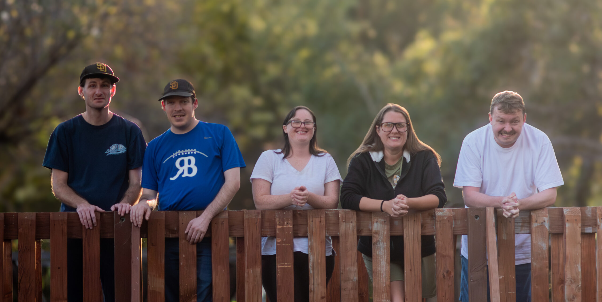 Five smiling Big Moves clients leaning on a wooden fence outdoors at golden hour