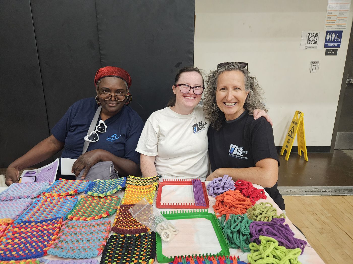 Members of the Big Moves community behind a table of handmade woven potholders at a community event